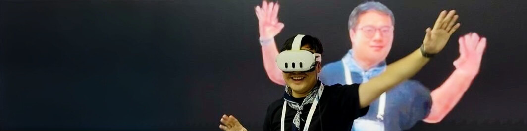 A person wearing a VR headset extends both arms in front of two large projected figures on a dark background.